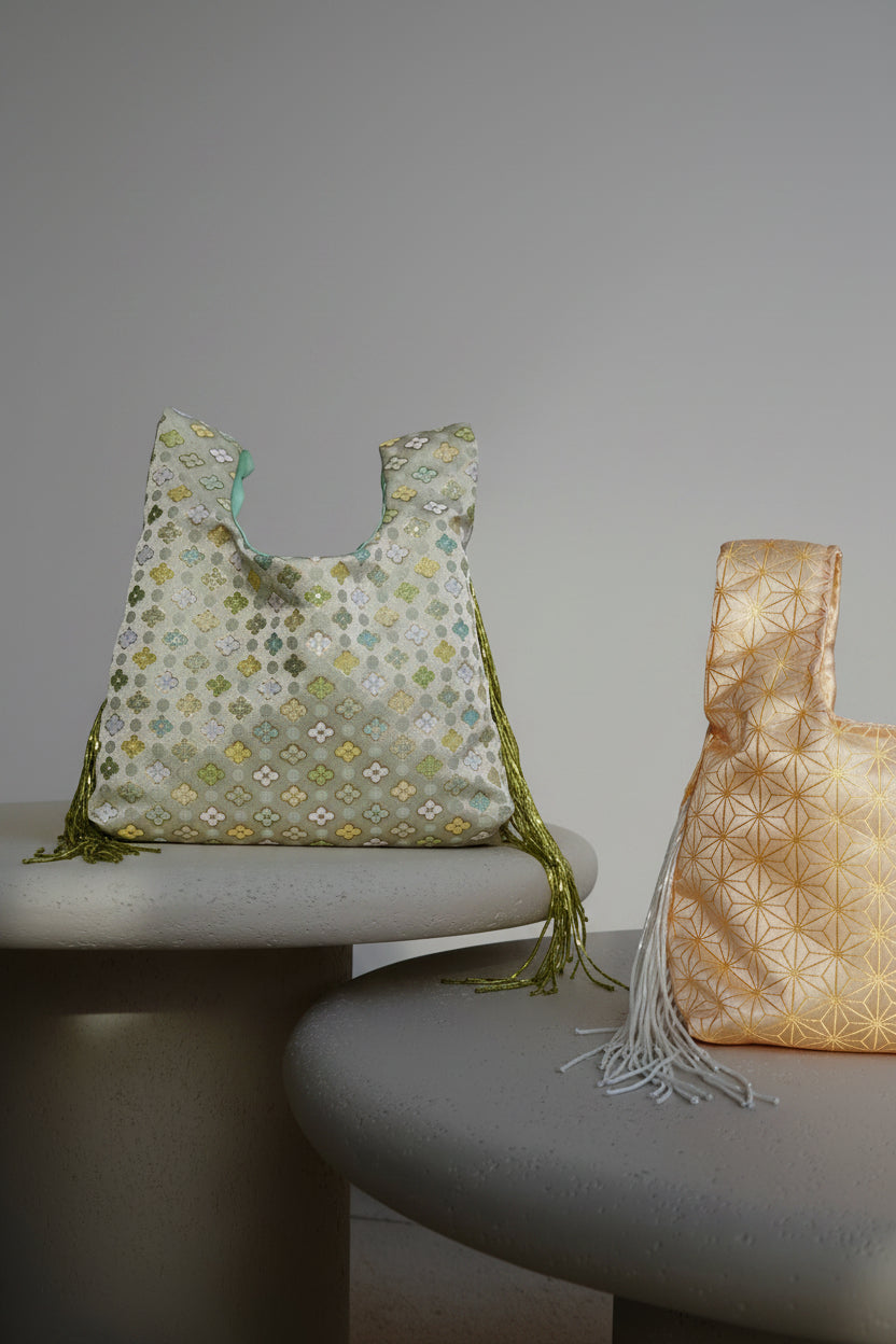 Two reusable tote bags with geometric patterns, one in sage green and white with olive green tassels, and one in pale gold and white with white tassels, displayed on a modern sofa in a sunlit room.