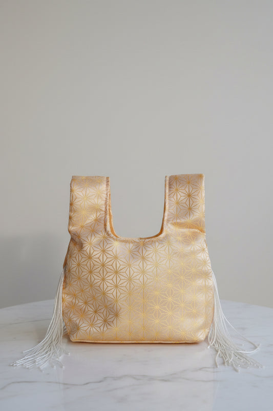 A small metallic gold handbag with a geometric star pattern and white fringe tassels, resting on a round gray table.