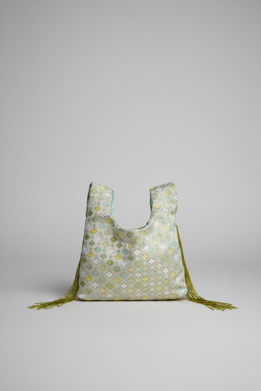 A light sage green reusable tote bag with a yellow and white floral pattern, featuring olive green tassels, resting on a white stone table in front of a cream-colored sofa.