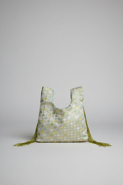 A light sage green reusable tote bag with a yellow and white floral pattern, featuring olive green tassels, resting on a white stone table in front of a cream-colored sofa.