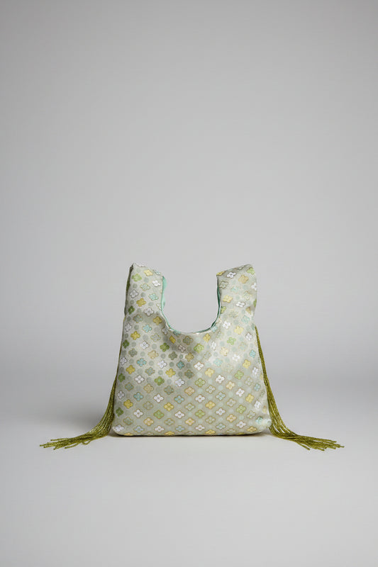 A light sage green reusable tote bag with a yellow and white floral pattern, featuring olive green tassels, resting on a white stone table in front of a cream-colored sofa.